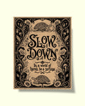 A square decorative art print or poster with ornate black floral scrollwork and the phrase 'SLOW DOWN In a world of hares, be a tortoise' on a warm beige background, shown against a light cream backdrop.